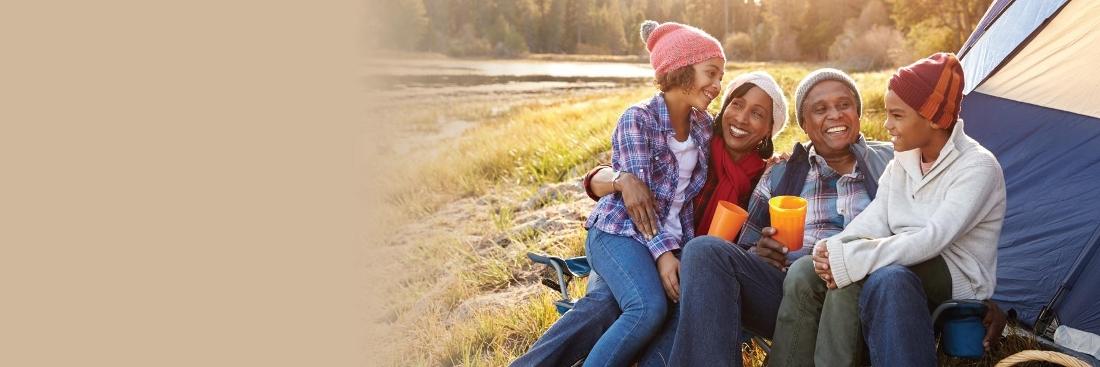 family of four cuddling up between a lake and a tent wearing chilly weather clothing and hats